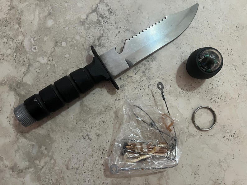 May include: A survival knife with a serrated blade, black handle, and a silver-colored blade. Includes a compass, a small metal ring, and a small survival kit in a clear plastic bag.