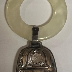 May include: A vintage silver bell with a cream-colored ring handle. The bell features a clock face and an inscription with the date "October 26, 1951". The bell is a unique collectible item.