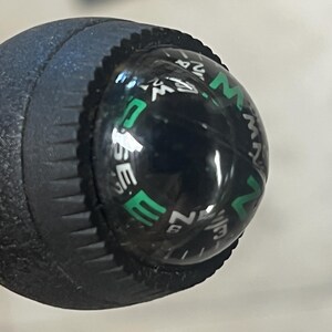 May include: Close-up of a black compass with a clear, rounded top. The compass rose is visible with white and green lettering indicating directions. The compass is attached to a textured, dark blue handle.