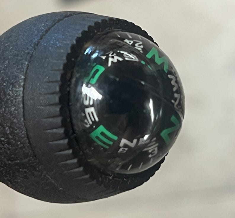 May include: Close-up of a black compass with a clear, rounded top. The compass rose is visible with white and green lettering indicating directions. The compass is attached to a textured, dark blue handle.