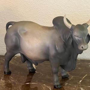 May include: A ceramic figurine of a bull with a matte grey finish, featuring a prominent hump on its back, a thick neck, and light-colored horns. The bull is depicted in a standing position with its head turned slightly toward the right side.