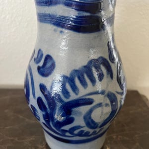 May include: A small, light blue ceramic pitcher with a rounded body and a slightly flared rim. The pitcher is decorated with dark blue floral and swirl patterns. The rim features horizontal dark blue brushstrokes. The pitcher sits on a dark brown surface.
