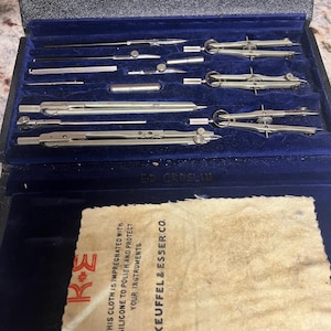 May include: A vintage drafting set in a black case with a blue velvet interior. The set includes various metal compasses, dividers, and other drawing tools. A cleaning cloth with the text "KEUFFEL & ESSER CO." is also included.