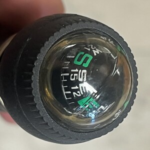 May include: Close-up of a black compass with a clear, domed top. The compass dial features green and white markings, including the letters "S" and "SE", and numbers. The compass is encased in a textured black rubber grip.