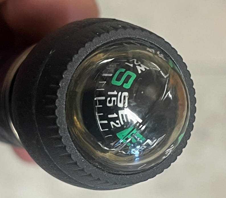 May include: Close-up of a black compass with a clear, domed top. The compass dial features green and white markings, including the letters "S" and "SE", and numbers. The compass is encased in a textured black rubber grip.