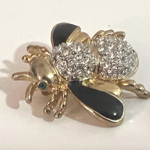 May include: A gold-toned bee brooch featuring black enamel wings, a body covered in clear sparkling rhinestones, and a small green rhinestone eye. The insect-shaped pin has a polished metallic finish and detailed legs and antennae.