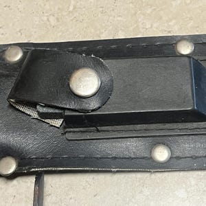 May include: Close-up of a black leather sheath with silver rivets and a black strap secured by a silver snap. The sheath has a rectangular black holder and a small piece of mesh.