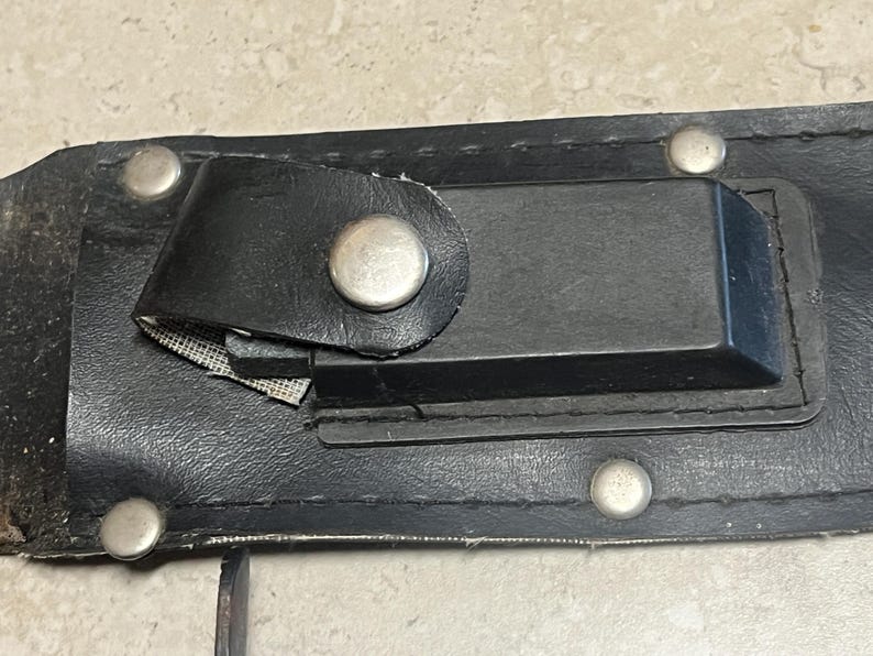 May include: Close-up of a black leather sheath with silver rivets and a black strap secured by a silver snap. The sheath has a rectangular black holder and a small piece of mesh.