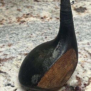 Pu&ograve; includere: Un'antica mazza da golf con testa nera e faccia in legno. La testa della mazza &egrave; di colore scuro, con una sezione in legno marrone pi&ugrave; chiara. L'impugnatura &egrave; avvolta con nastro nero. Un articolo sportivo vintage.