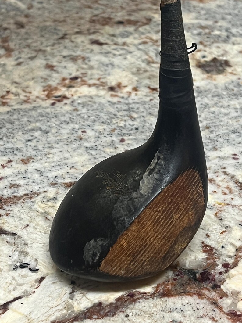 Pu&ograve; includere: Un'antica mazza da golf con testa nera e faccia in legno. La testa della mazza &egrave; di colore scuro, con una sezione in legno marrone pi&ugrave; chiara. L'impugnatura &egrave; avvolta con nastro nero. Un articolo sportivo vintage.