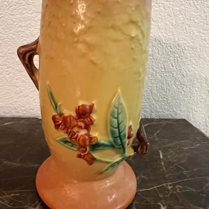 Puede incluir: Un jarrón de cerámica vintage con un acabado amarillo-naranja texturizado. El jarrón presenta un diseño floral con detalles en rojo y verde, y un asa marrón. La base es de color naranja claro. Una pieza decorativa para la decoración del hogar.