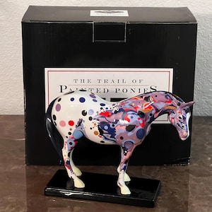 May include: A decorative horse figurine with a colorful abstract design. The horse is white with blue, red, purple, and gold spots and patterns. It stands on a black rectangular base, with the words "THE TRAIL OF PAINTED PONIES" on the box behind it.