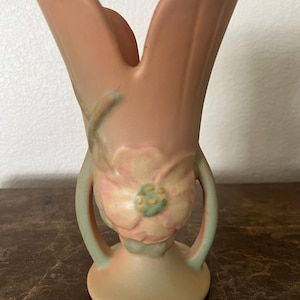 May include: A small, pink and green ceramic vase with a floral design. The vase has two handles and a wide base.