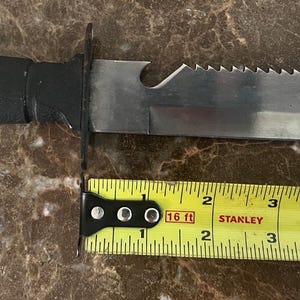 May include: A black-handled knife with a serrated blade and a partially serrated spine. The blade is approximately 6 inches long. A yellow tape measure is placed next to the knife.
