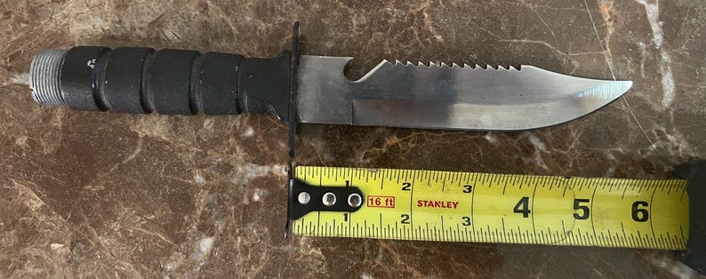 May include: A black-handled knife with a serrated blade and a partially serrated spine. The blade is approximately 6 inches long. A yellow tape measure is placed next to the knife.