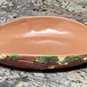 May include: A peach-colored ceramic serving dish with a scalloped rim and handles. The dish is decorated with a relief pattern of white flowers and green leaves along the bottom edge. A vintage piece.