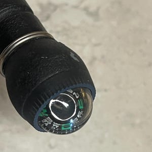 May include: Close-up of a black compass with a clear, rounded top. The compass dial features white and green markings, including directional indicators. A silver metal ring encircles the compass and the black handle.