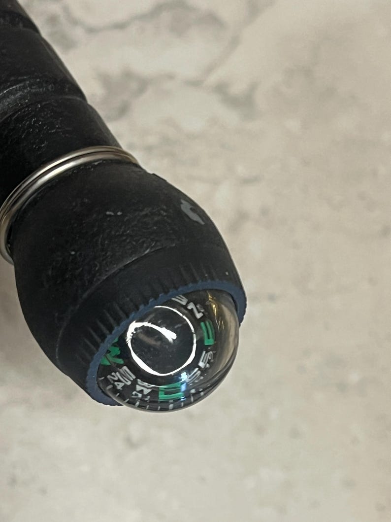May include: Close-up of a black compass with a clear, rounded top. The compass dial features white and green markings, including directional indicators. A silver metal ring encircles the compass and the black handle.