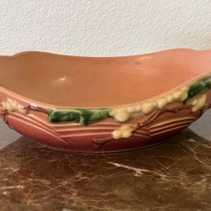 May include: A peach-colored ceramic bowl with a scalloped rim and handles. The bowl features a decorative relief design with green and yellow accents. The bowl is oval-shaped and sits on a marble surface.