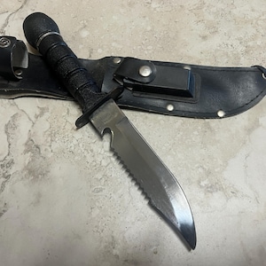 May include: A black survival knife with a serrated blade and a black handle, resting on a black leather sheath. The knife has a sturdy design, suitable for outdoor activities. The sheath has a belt loop and a snap closure.