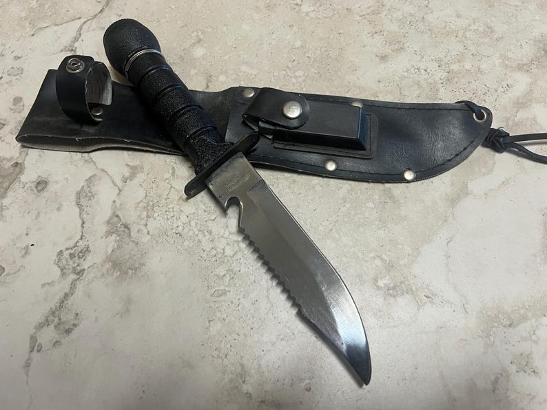 May include: A black survival knife with a serrated blade and a black handle, resting on a black leather sheath. The knife has a sturdy design, suitable for outdoor activities. The sheath has a belt loop and a snap closure.
