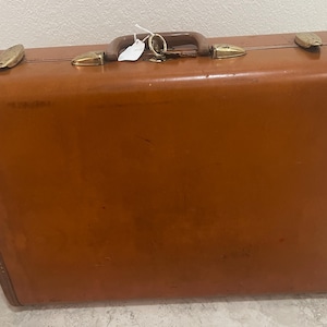 May include: A vintage brown suitcase with brass-colored hardware. The suitcase has a leather handle and corner protectors. The exterior is a solid, glossy brown with a few minor scuffs. The suitcase is rectangular and appears to be in good condition.
