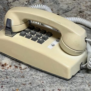 May include: Cream-colored vintage telephone with a handset and a coiled cord. The phone has a keypad with dark gray buttons and a silver metal accent. The phone is sitting on a speckled surface.