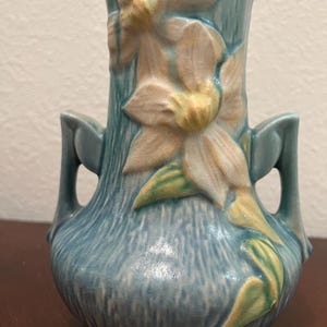 May include: A blue ceramic vase with a floral design. The vase features a textured surface and two handles. The flowers are cream and yellow, with green leaves. The vase is approximately 8 inches tall.