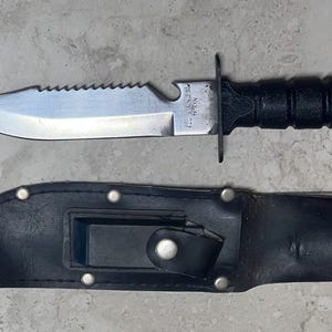 May include: A black and silver knife with a serrated edge and a black handle, accompanied by a black leather sheath. The knife has a pointed blade and a guard. The sheath has a snap closure and a belt loop.