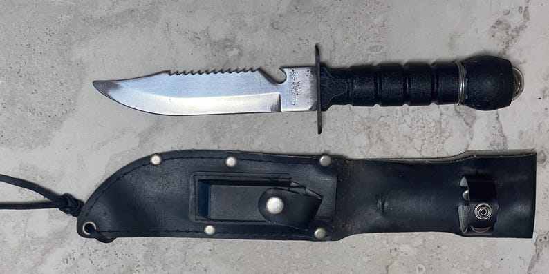 May include: A black and silver knife with a serrated edge and a black handle, accompanied by a black leather sheath. The knife has a pointed blade and a guard. The sheath has a snap closure and a belt loop.