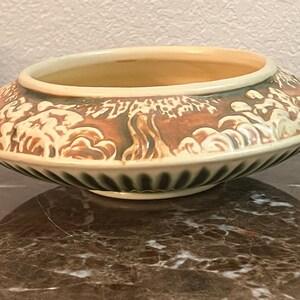 May include: A low, round ceramic bowl with a wide rim. The bowl features a detailed relief design of trees and figures in shades of brown and cream against a green and cream background. The interior is a light tan color.