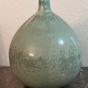May include: A round ceramic vase with a narrow neck, featuring a mottled sage green glaze with lighter, speckled white patterns across the surface. The finish has a smooth, glossy texture that creates subtle variations in color and light reflection.