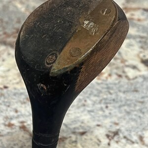 Pu&ograve; includere: Un'antica mazza da golf con una testa in legno scuro e una piastra metallica. La testa della mazza mostra segni di usura e invecchiamento, con un'impugnatura nera e un aspetto usurato. La mazza &egrave; probabilmente vintage.