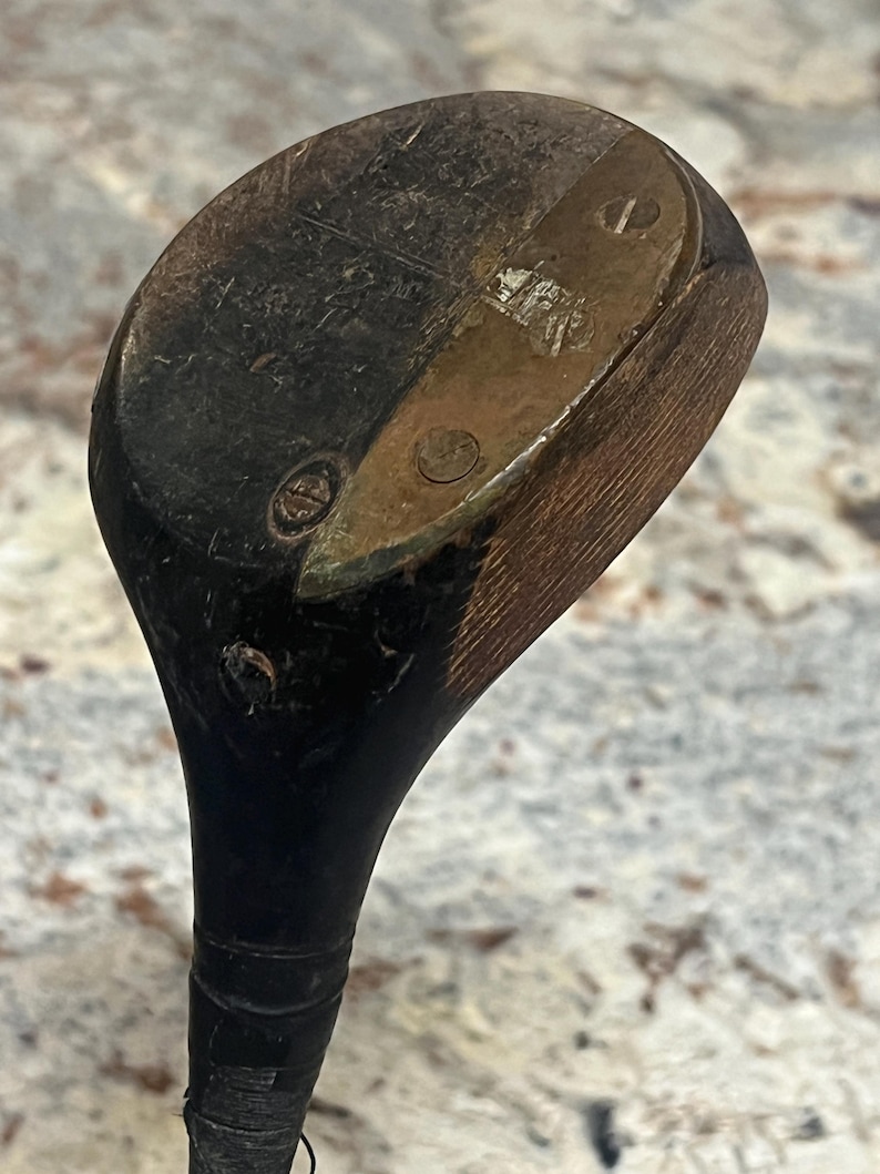 Pu&ograve; includere: Un'antica mazza da golf con una testa in legno scuro e una piastra metallica. La testa della mazza mostra segni di usura e invecchiamento, con un'impugnatura nera e un aspetto usurato. La mazza &egrave; probabilmente vintage.