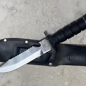 May include: A black knife with a serrated blade is partially inserted into a black leather sheath. The knife has a black handle and a silver blade. The sheath has a loop and a snap closure. The knife is approximately 10 inches long.