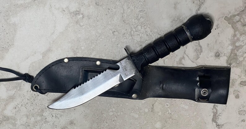 May include: A black knife with a serrated blade is partially inserted into a black leather sheath. The knife has a black handle and a silver blade. The sheath has a loop and a snap closure. The knife is approximately 10 inches long.