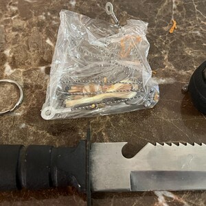 May include: Survival kit with a black-handled knife, a compass, a metal keyring, and a small bag of survival tools. The knife has a serrated edge and a silver blade. The compass is black with a clear top. The kit is on a brown marble surface.