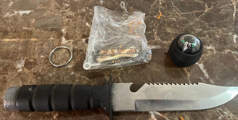 May include: Survival kit with a black-handled knife, a compass, a metal keyring, and a small bag of survival tools. The knife has a serrated edge and a silver blade. The compass is black with a clear top. The kit is on a brown marble surface.
