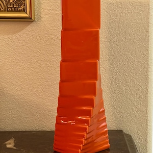 May include: A tall, bright orange ceramic vase with a distinct geometric design. The structure features a series of stacked, rectangular blocks that create a stepped, tapering silhouette from the wider base up to the narrower top section.