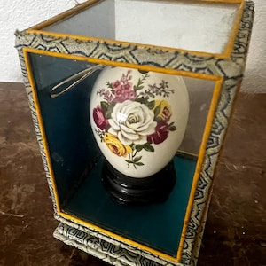 May include: A white porcelain egg with a floral design is displayed in a glass case with a yellow and green patterned base. The egg is on a black stand and has a gold string attached to the top.