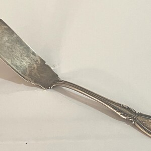 May include: A silver butter knife with a curved, serrated blade and a decorative handle. The handle has an ornate design with a small, engraved initial. The knife is made of a shiny, reflective metal.
