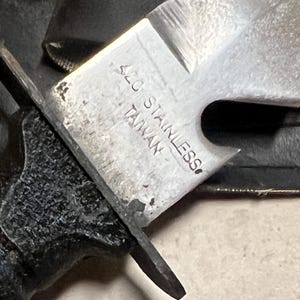 May include: Close-up of a stainless steel knife blade with a partially visible handle. The blade has a serrated edge and the text "420 STAINLESS TAIWAN" is stamped on the metal. The handle is black and textured.