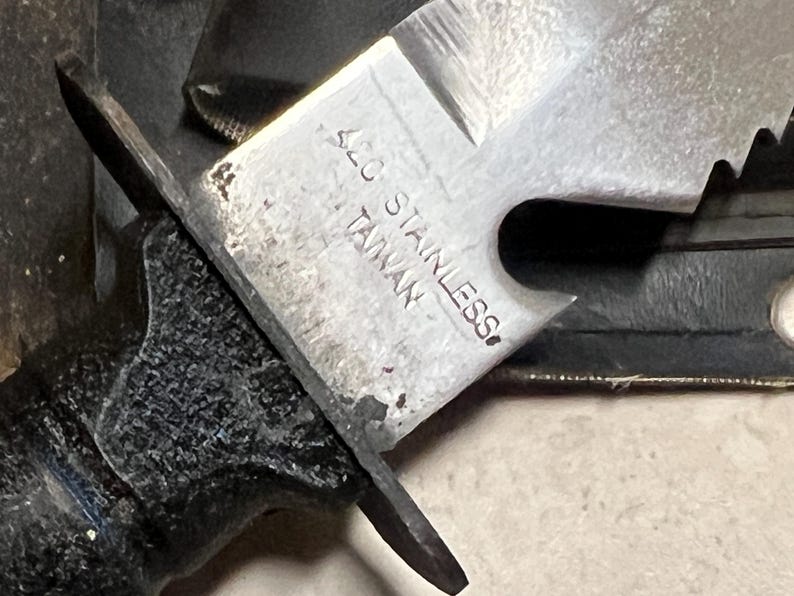 May include: Close-up of a stainless steel knife blade with a partially visible handle. The blade has a serrated edge and the text "420 STAINLESS TAIWAN" is stamped on the metal. The handle is black and textured.