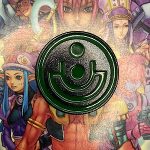 May include: A green metal pin with a stylized design of a circle with a smaller circle inside and a curved line underneath. The pin is surrounded by a colorful cartoon image of characters from a video game.