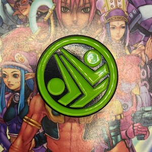May include: A round, green and black enamel pin featuring a stylized design with a green circle and a geometric shape. The pin is set against a background of anime-style characters in various colors.