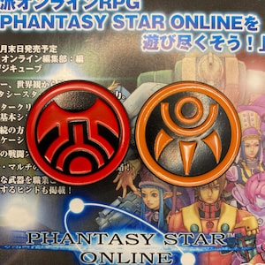 Op de afbeelding: Twee rode en oranje metalen pinnen met zwart-witte ontwerpen. De pinnen zijn van het videospel Phantasy Star Online. De tekst "Phantasy Star Online" staat onderaan de afbeelding.