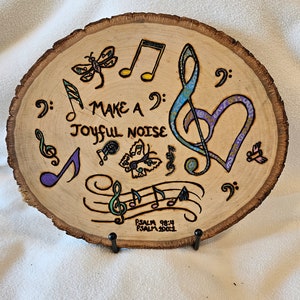 May include: A wooden round slice with a wood burned design of musical notes, a treble clef, and butterflies. The text "Make a Joyful Noise" is written in cursive. The text "Psalm 98:4 Psalm 100:1" is written in smaller font below the design.