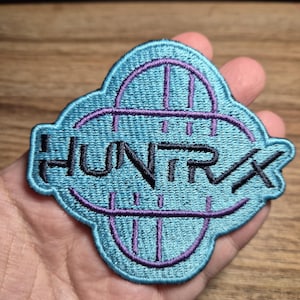 May include: A teal embroidered patch with the word "HUNTRX" in black, framed by a purple arched design. The patch is held in a hand, with a wooden surface in the background.