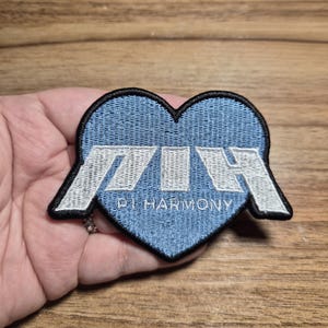 May include: A heart-shaped patch with a light blue background and the text "TIH PI HARMONY" in white. The patch has a black border and is likely an embroidered or fabric item.