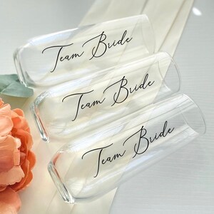 May include: Three clear glass champagne flutes with the text "Team Bride" in black calligraphy on each.